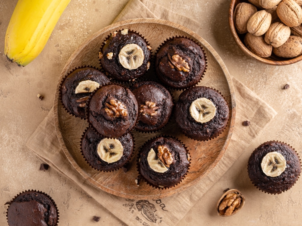 Chocolate banana muffins decorated with walnuts and banana slices on wooden plate.